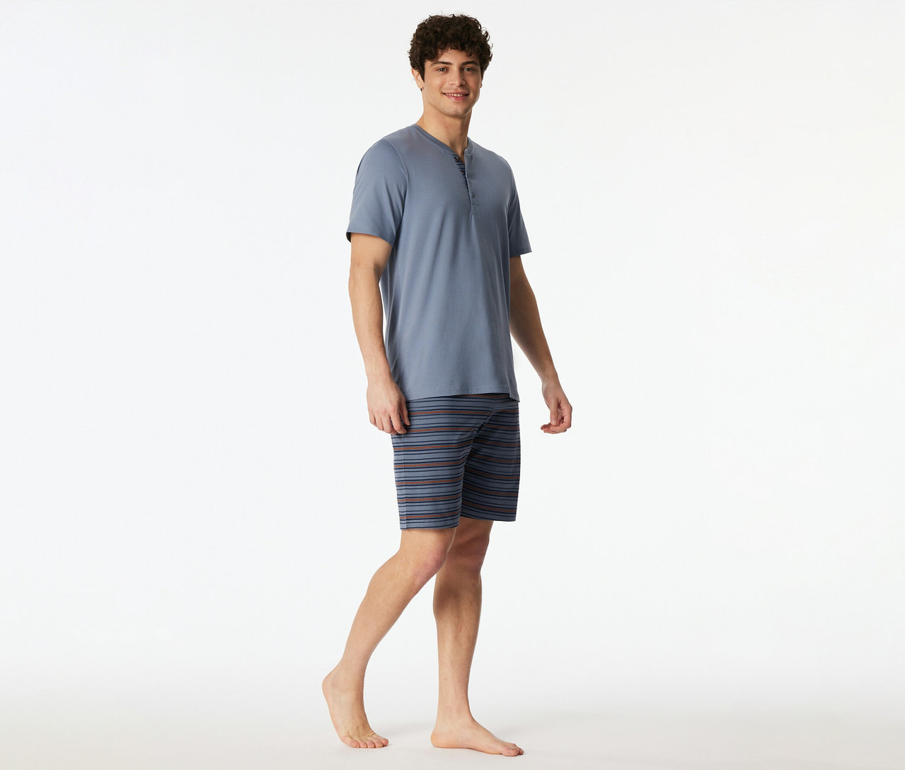 Un homme souriant porte un set shorty SCHIESSER bleu, composé d'un haut Henley à manches courtes et d'un short rayé, debout pieds nus.