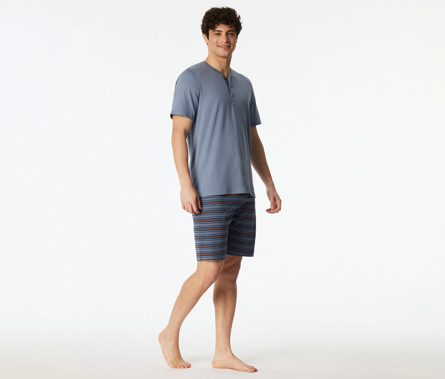 Un homme souriant porte un set shorty SCHIESSER bleu, composé d'un haut Henley à manches courtes et d'un short rayé, debout pieds nus.