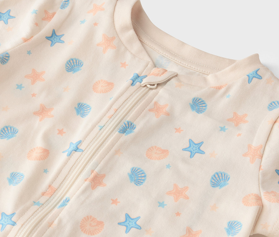 Gros plan d'une grenouillère pour bébé de couleur crème avec fermeture éclair et motif intégral de coquillages bleus et d'étoiles de mer couleur pêche.