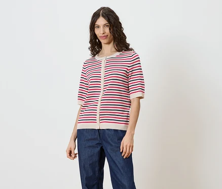 Une femme aux cheveux bruns ondulés porte un cardigan rayé à manches courtes boutonné et un jean sur fond blanc.