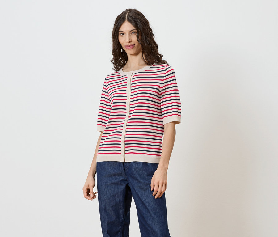 Une femme aux cheveux bruns ondulés porte un cardigan rayé à manches courtes boutonné et un jean sur fond blanc.