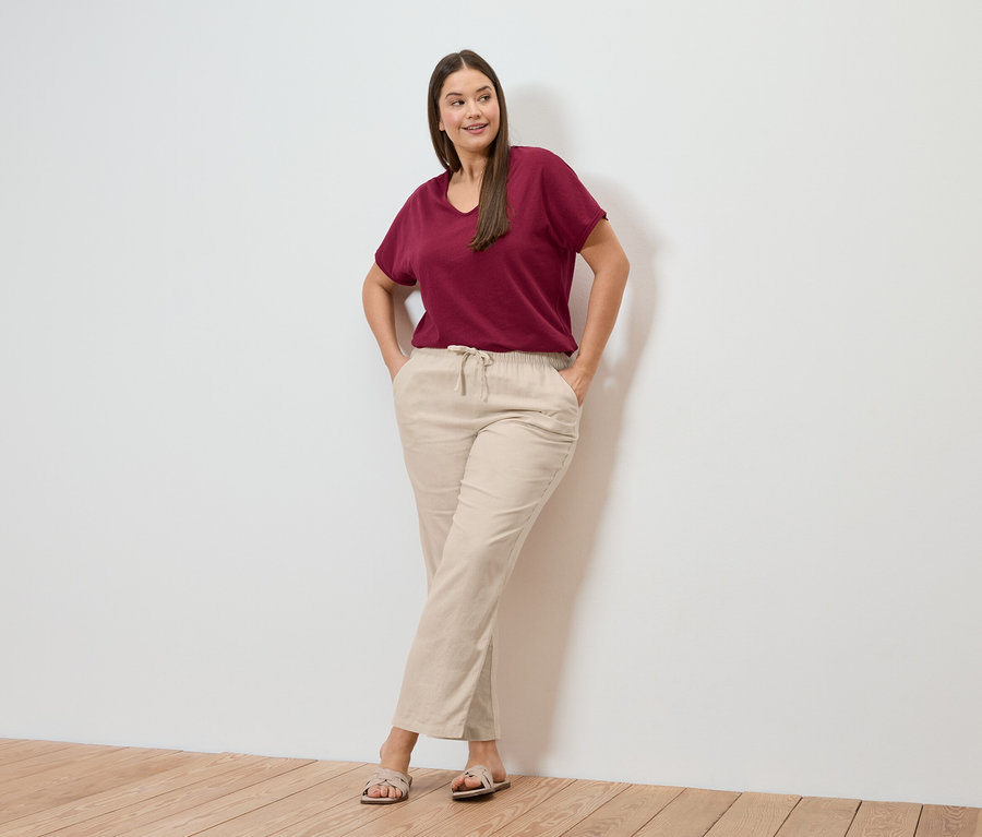 Une femme pose devant un mur, portant un t-shirt rouge foncé en lin mélangé et un pantalon en lin crème.