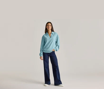 Femme portant un haut bleu clair à manches longues avec un col et un jean foncé, une main dans la poche.