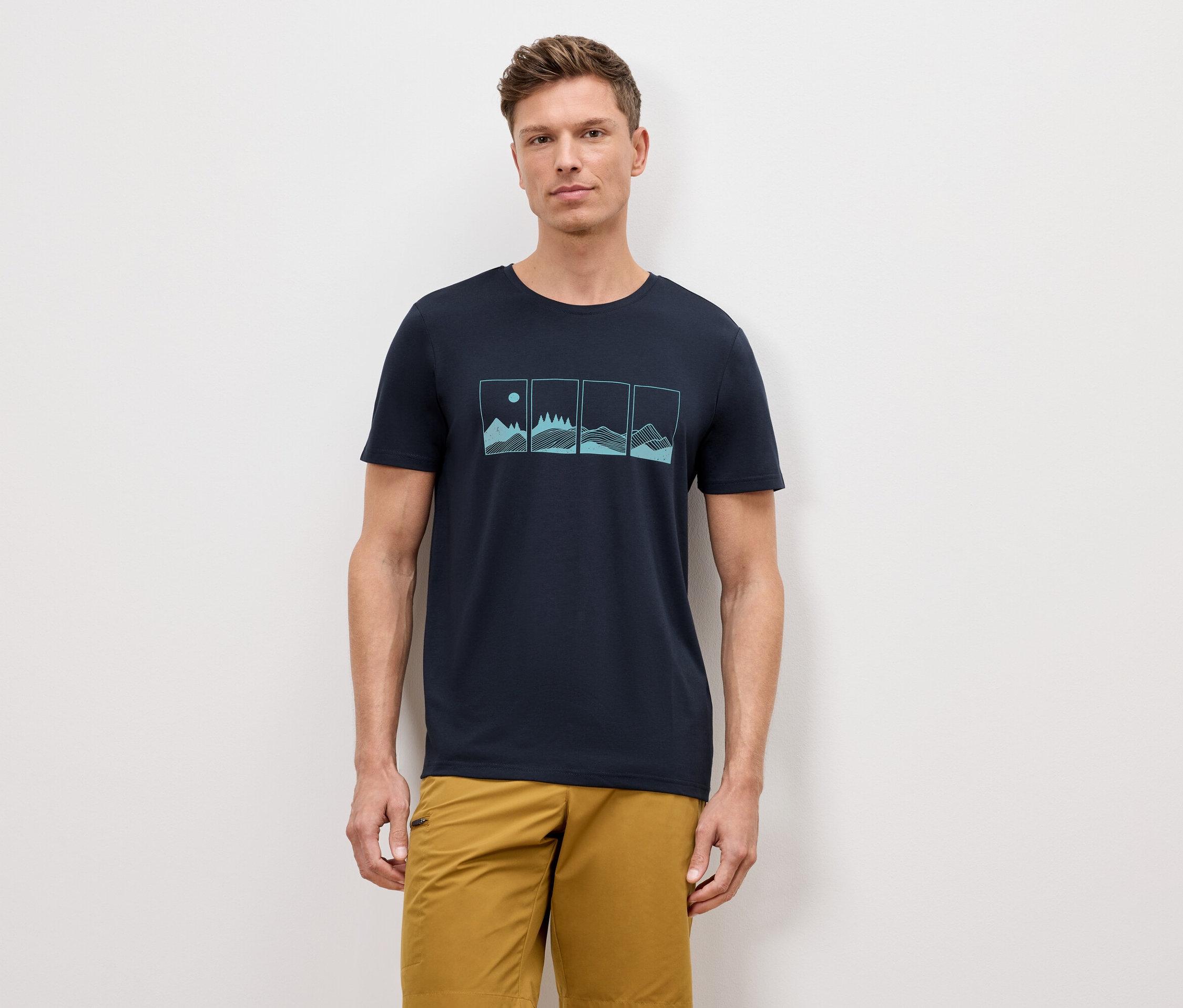 Un homme se tient devant un mur, portant un t-shirt bleu marine avec un motif de quatre paysages naturels et un short kaki.