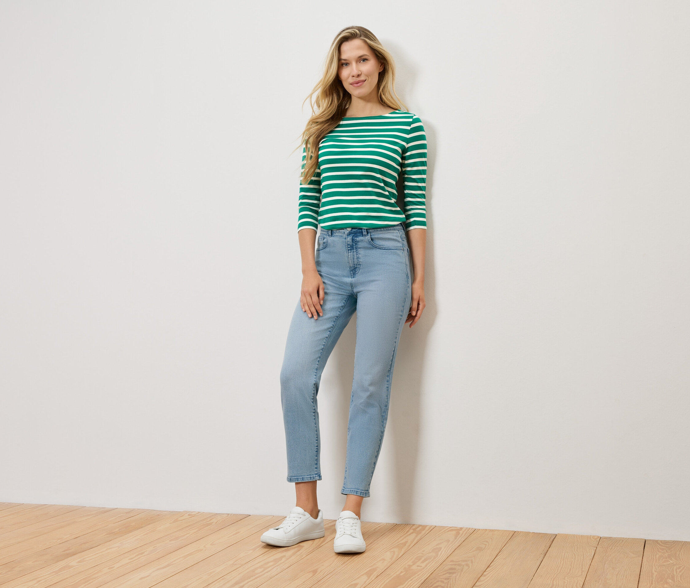 Une femme blonde se tient devant un mur, portant un t-shirt à manches longues vert et blanc rayé, un jean bleu clair et des baskets blanches.