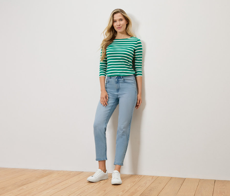 Une femme blonde se tient devant un mur, portant un t-shirt à manches longues vert et blanc rayé, un jean bleu clair et des baskets blanches.