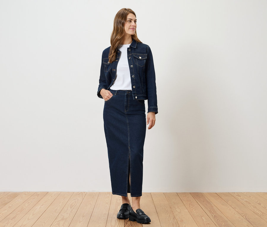 Une femme debout porte une veste en jean et une jupe en jean fendue.