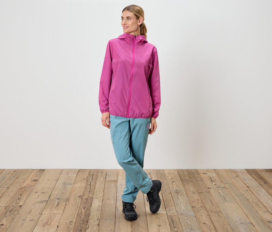 Une femme se tient debout, les jambes croisées, portant une veste rose, un pantalon bleu et des chaussures noires.