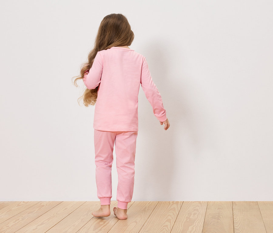 Fillette aux longs cheveux bruns, vue de dos, portant un pyjama rose devant un mur blanc.