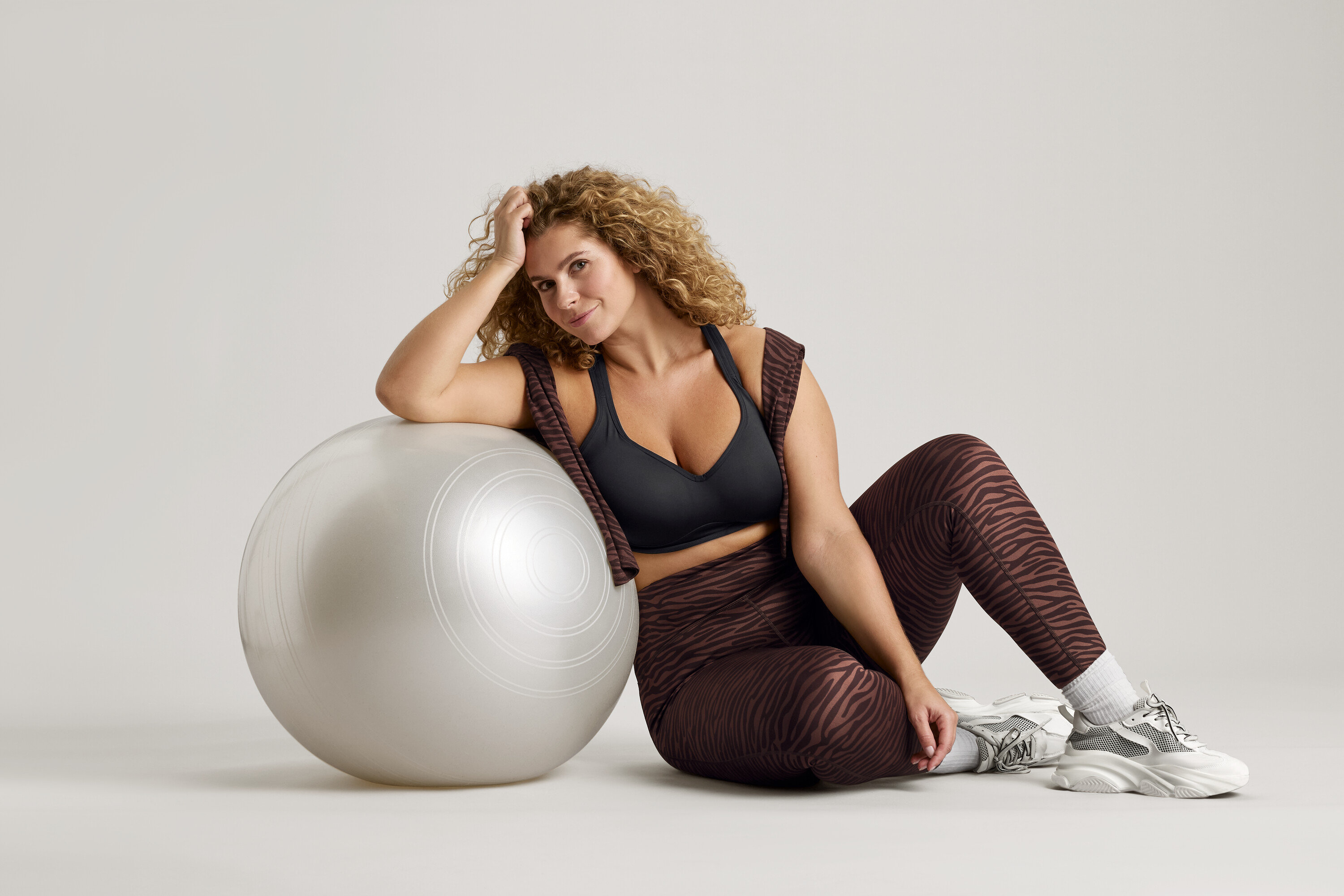 Femme aux cheveux bouclés assise à côté d'un ballon de gymnastique argenté, portant un soutien-gorge de sport noir, un legging à motif zébré et des baskets.