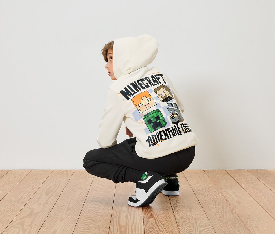 Un garçon est accroupi sur un plancher en bois, portant un sweat à capuche Minecraft de couleur crème et un pantalon noir.