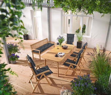 Sur une terrasse, une table de salle à manger en teck double extensible, un banc de jardin classique en teck, des chaises hautes en teck avec des coussins de dossier anthracite, un pot de fleurs avec un tissage textile et une lampe de table solaire rechargeable grise.