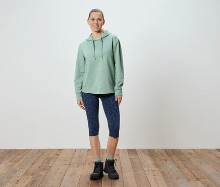 Une femme se tient debout, portant un sweat à capuche vert clair, un legging Capri bleu à motifs et des chaussures de randonnée noires.