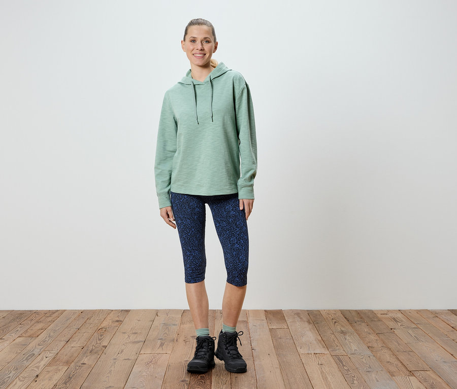 Une femme se tient debout, portant un sweat à capuche vert clair, un legging Capri bleu à motifs et des chaussures de randonnée noires.