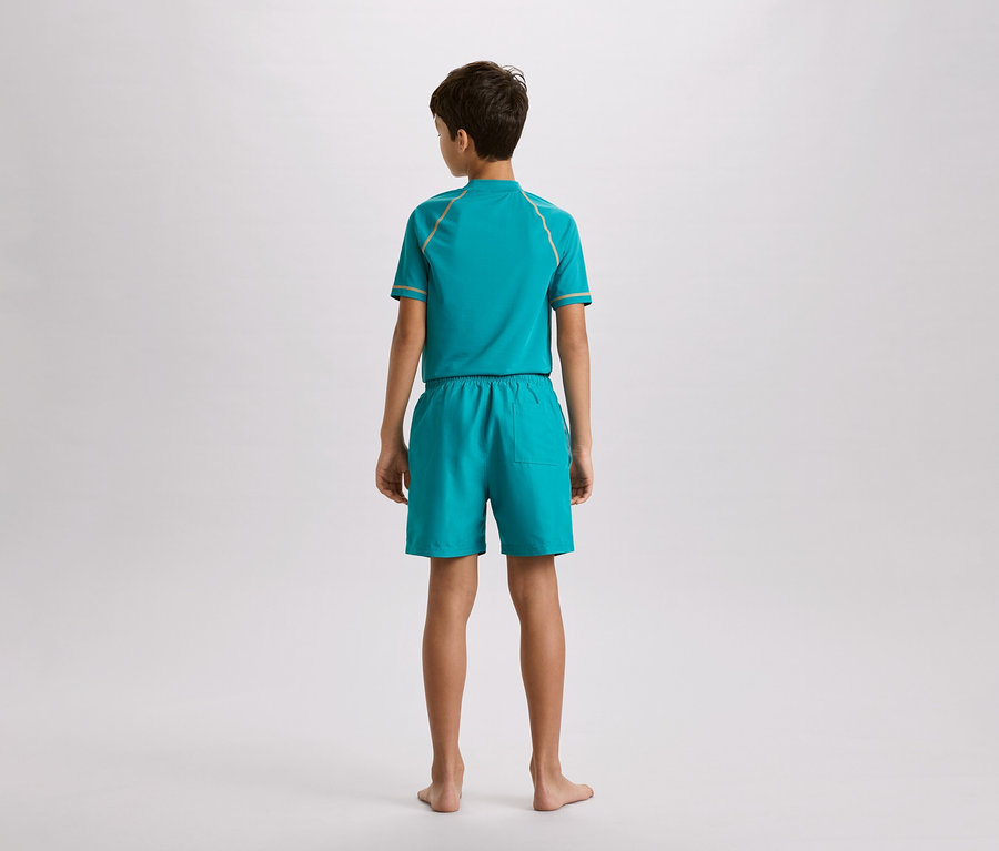 Garçon vu de dos, portant un ensemble turquoise composé d'un haut et d'un short avec des détails dorés, sur un fond neutre.