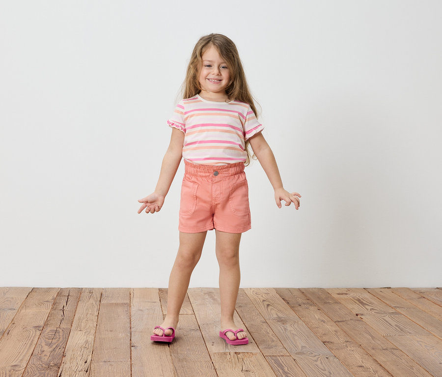 Fillette blonde aux cheveux longs, portant un t-shirt rayé, un short et des sandales.