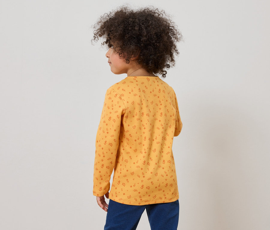 Enfant aux cheveux bouclés portant un haut jaune à manches longues avec un motif floral et un pantalon bleu, vu de dos.