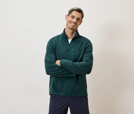 Un homme souriant, les bras croisés, porte une veste en polaire verte zippée et un short bleu marine sur fond clair.