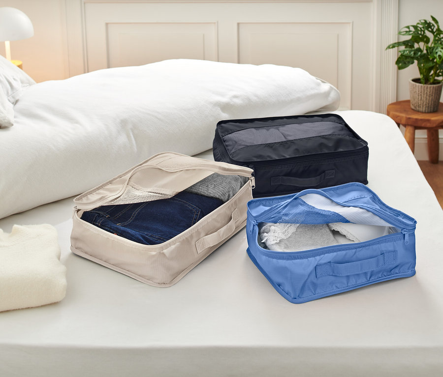 Trois cubes de rangement ouverts sont posés sur un lit avec des draps blancs. Les cubes de rangement sont beiges, bleus et noirs et contiennent des vêtements.