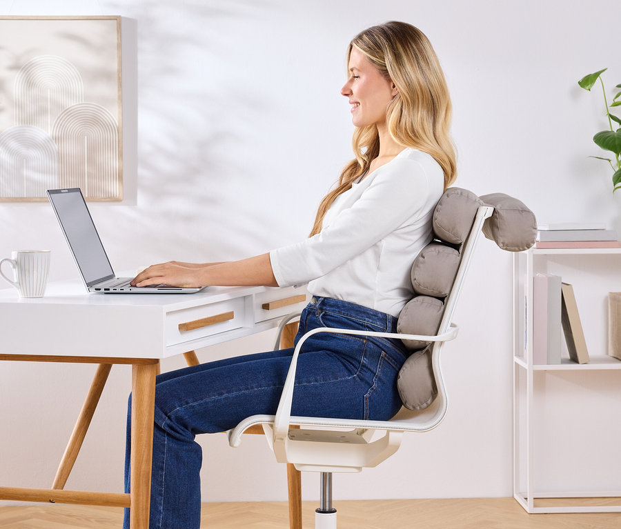 Une femme est assise à un bureau et utilise un ordinateur portable, avec un coussin de soutien dorsal gris sur sa chaise.
