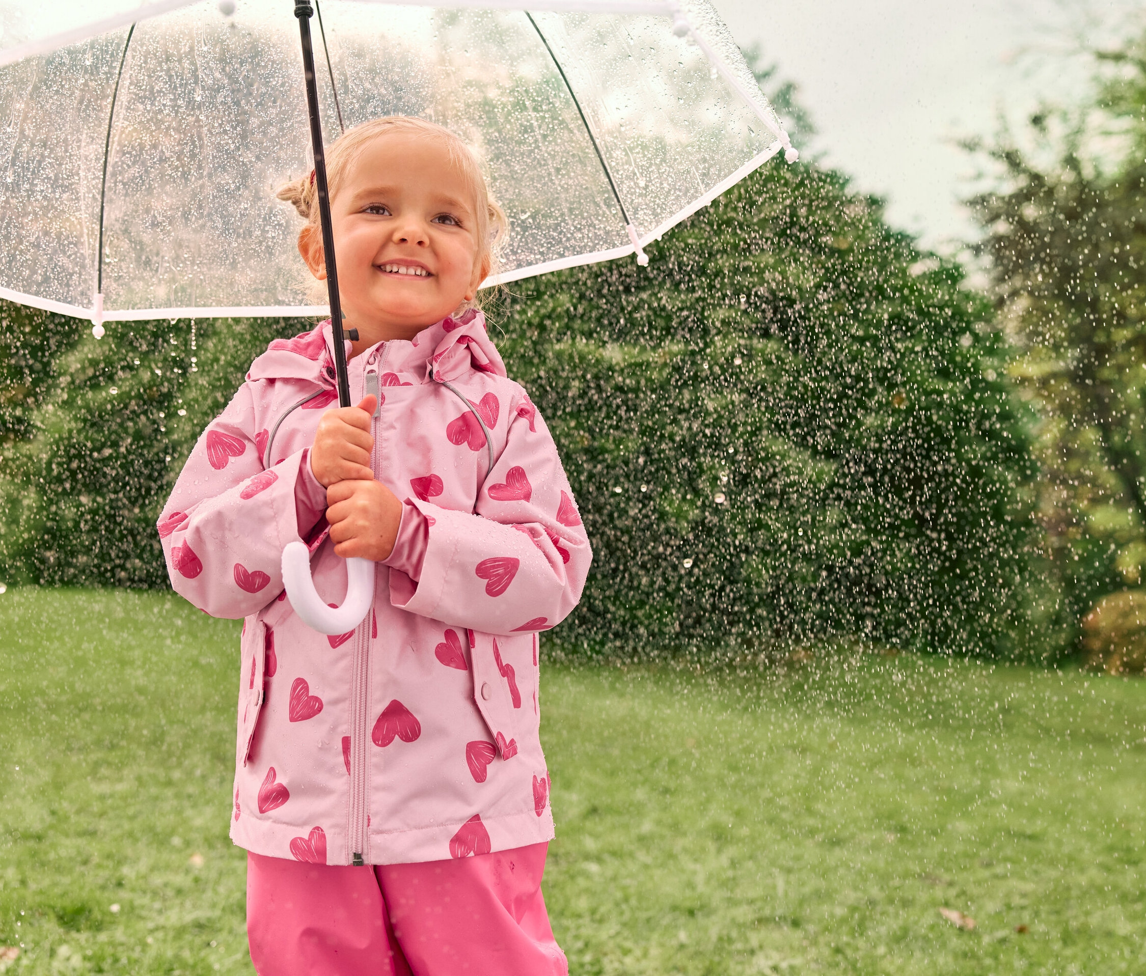 Une petite fille blonde sourit sous un parapluie transparent sous la pluie, vêtue d'une veste rose à motifs de cœurs.