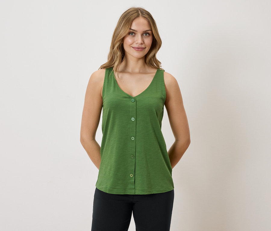 Une femme aux cheveux blonds porte un débardeur vert boutonné et un pantalon noir sur un fond blanc.