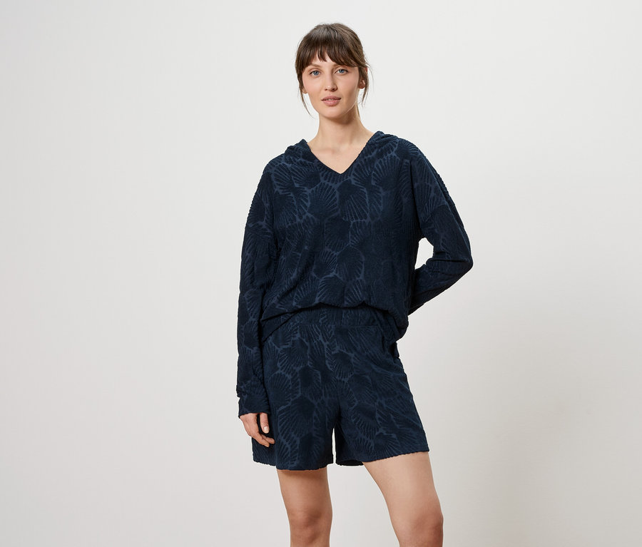 Une femme porte un haut à manches longues bleu marine à motifs avec un col en V et un short devant un fond neutre.
