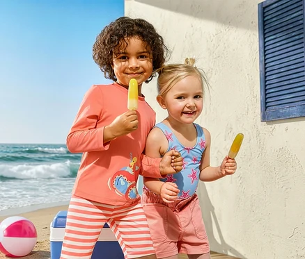 Deux enfants avec des glaces à la plage. La fille porte un maillot de bain pour enfant, étoiles de mer et un short en jean pour enfant. Le garçon porte un ensemble pour enfant avec indice de protection UV 50+, poulpe.