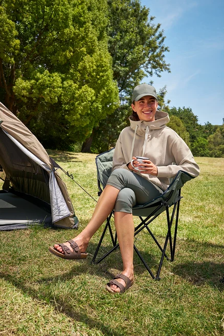 Une femme assise dans une chaise de camping Tambu »ASANA« tient une tasse et porte un legging 3/4 et un sweat à capuche.