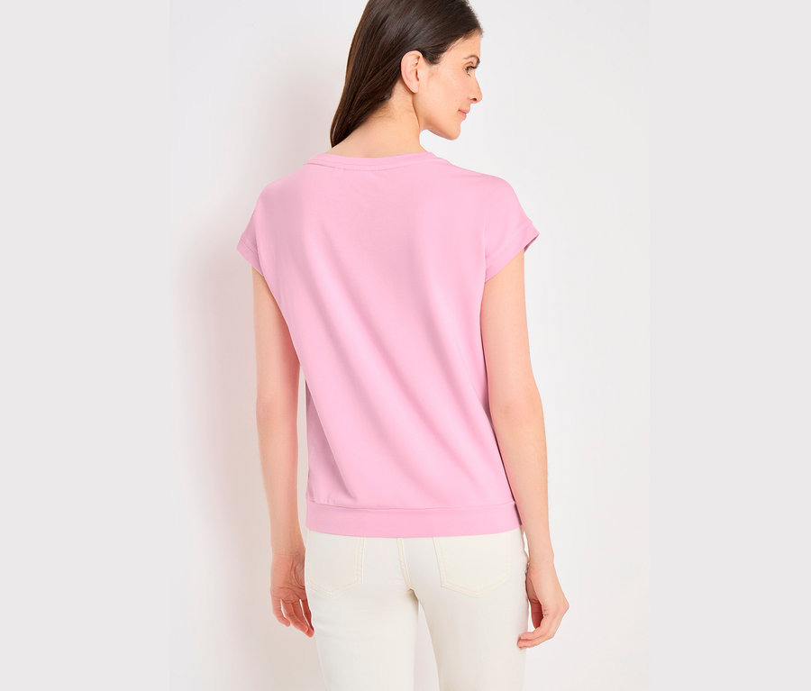 Femme vue de dos, portant un T-shirt à motif CECIL rose et un pantalon clair, regardant vers sa droite.