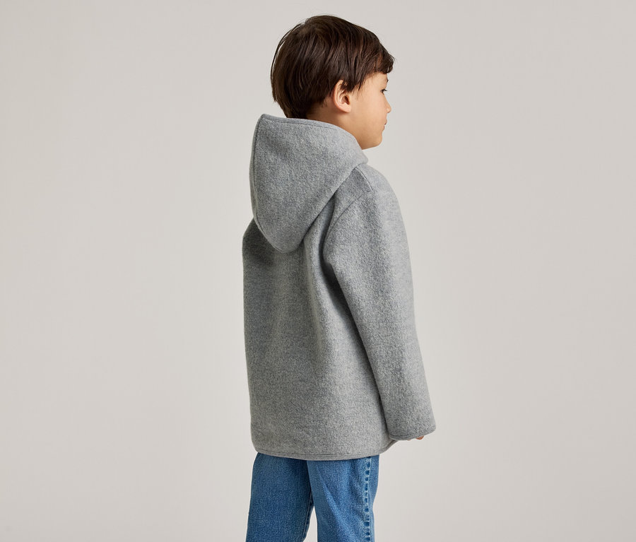 Un enfant aux cheveux bruns porte un manteau à capuche gris et un jean, vu de profil.