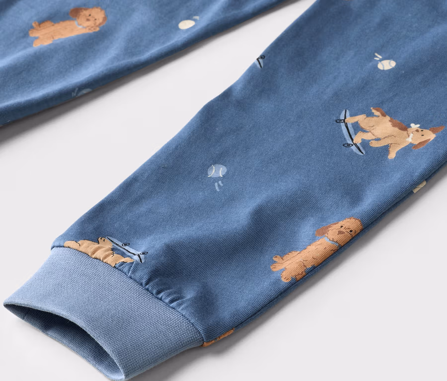 Gros plan d'un tissu bleu avec un motif de chiens de dessin animé marron et de petites balles de tennis blanches. Un chien est sur un skateboard. Le tissu a un poignet côtelé.