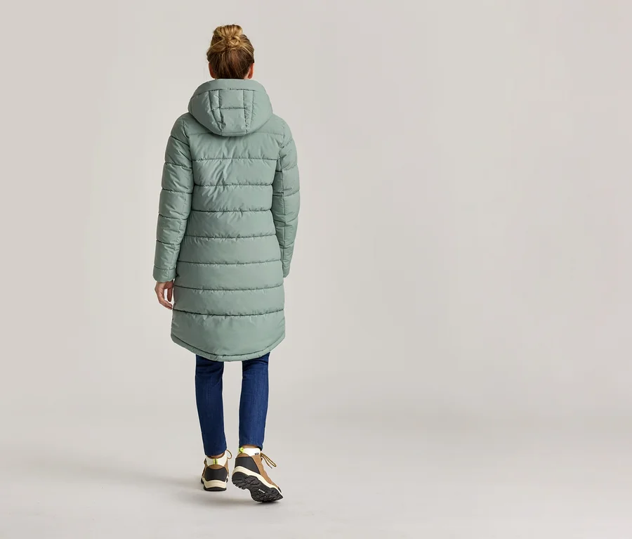 Vue arrière d'une femme portant un chignon, un long manteau matelassé vert menthe à capuche, un jean et des chaussures de randonnée.