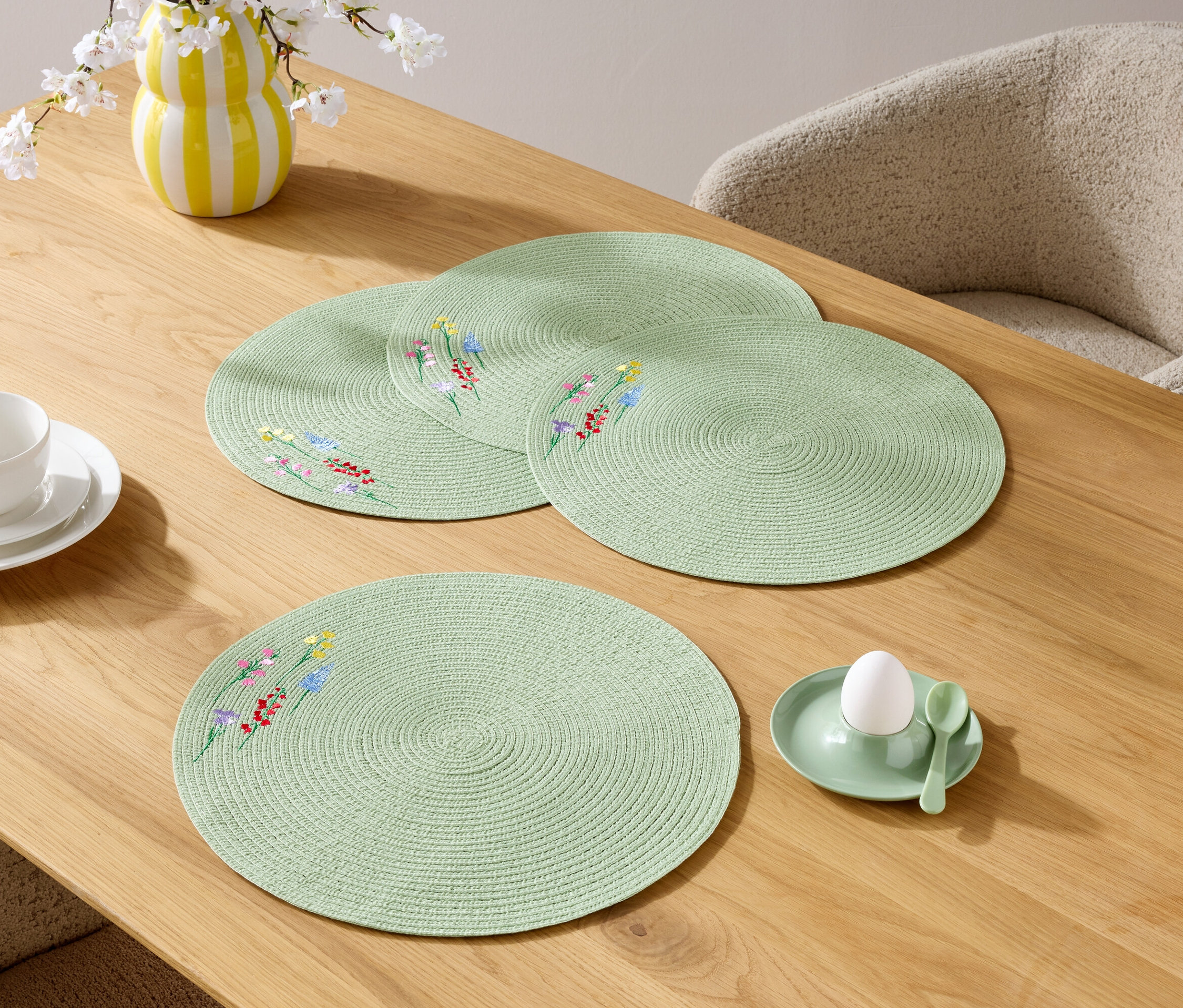 Gros plan de quatre sets de table ronds vert clair avec un motif floral sur une table en bois, ainsi qu'une tasse, une assiette, un vase de fleurs et un coquetier avec un œuf.