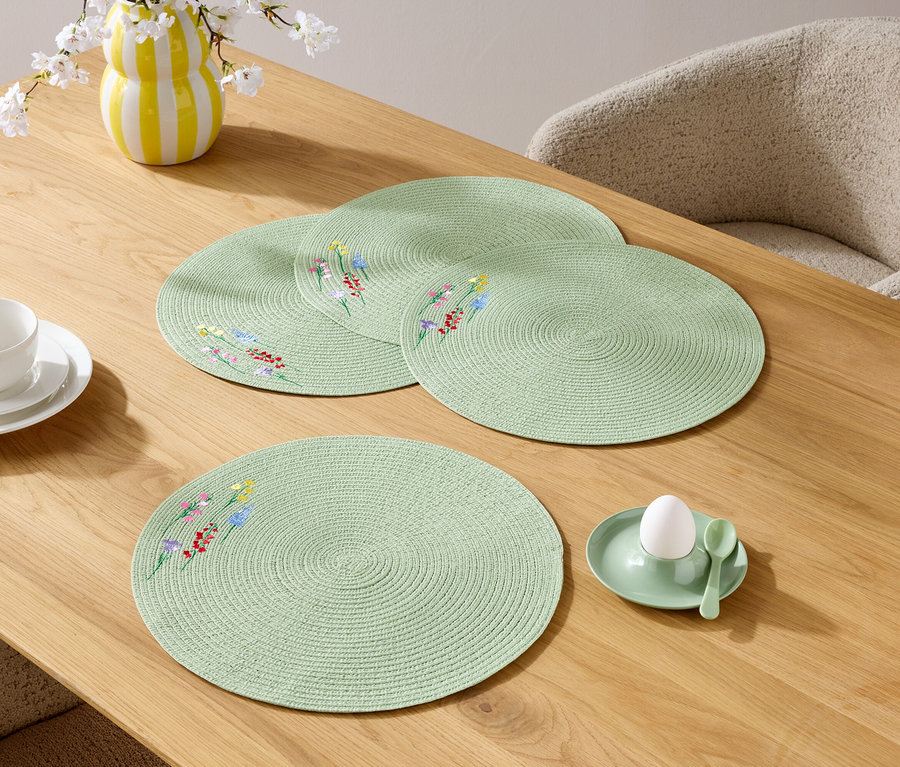 Gros plan de quatre sets de table ronds vert clair avec un motif floral sur une table en bois, ainsi qu'une tasse, une assiette, un vase de fleurs et un coquetier avec un œuf.