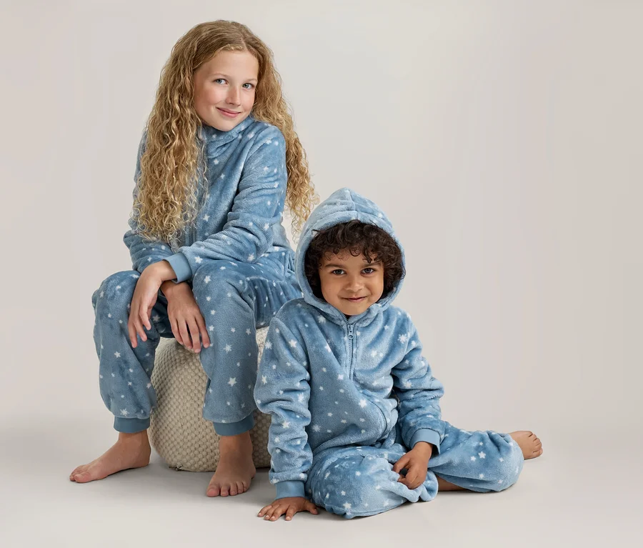 Deux enfants, une fille plus âgée et un jeune garçon, portent des pyjamas polaires bleus à capuche assortis avec des étoiles blanches. La fille est assise sur un pouf clair, le garçon est assis par terre, tous deux sourient.