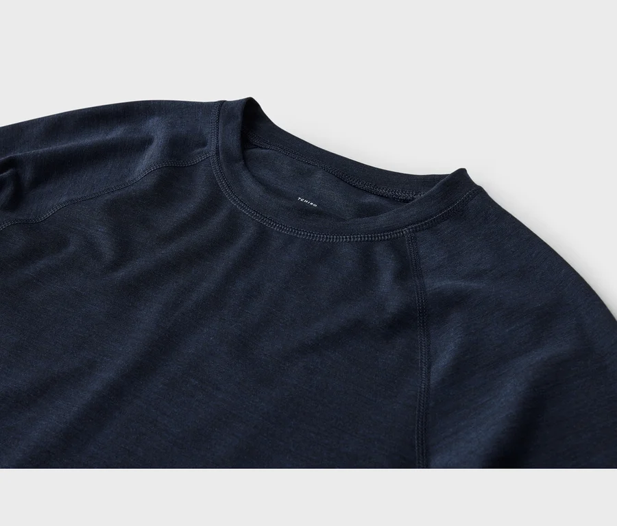 Gros plan d'un haut à manches longues bleu foncé avec un col rond et des manches raglan. Le tissu a une texture légèrement chinée, et des coutures visibles parcourent l'encolure et les coutures des manches. Une étiquette blanche « TCHIBO » est visible à l'intérieur du col.