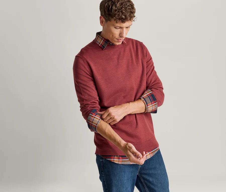 Homme remontant les manches de son pull rouge, révélant une chemise à carreaux en dessous, et portant un jean bleu.