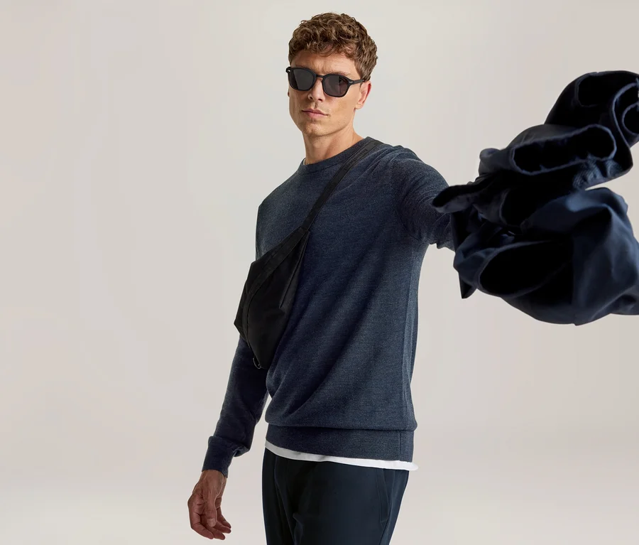 Homme en lunettes de soleil, pull bleu foncé et banane noire, tenant un vêtement bleu foncé froissé dans sa main tendue.