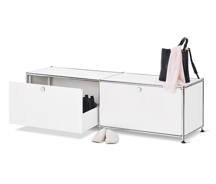 Un meuble bas en métal blanc « CN3 » ouvert avec des tiroirs et des chaussures à l'intérieur, à côté duquel se trouve une paire de chaussons crème, un sac à main noir et une écharpe rose sur le dessus du meuble.