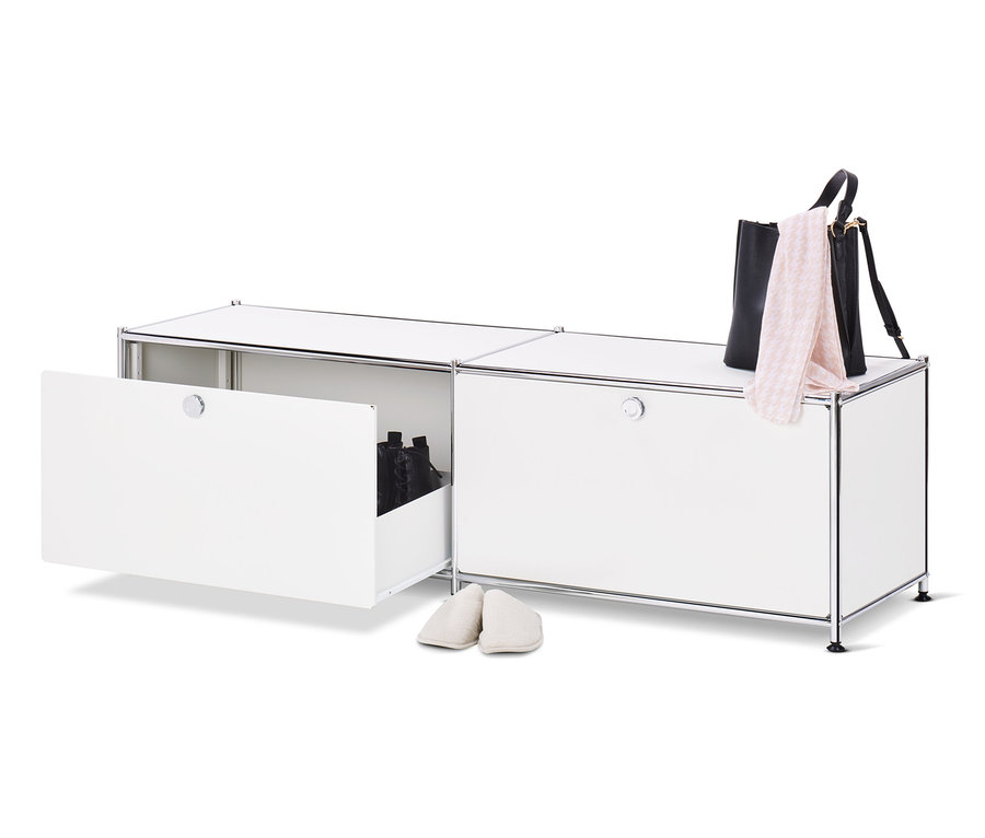 Un meuble bas en métal blanc « CN3 » ouvert avec des tiroirs et des chaussures à l'intérieur, à côté duquel se trouve une paire de chaussons crème, un sac à main noir et une écharpe rose sur le dessus du meuble.
