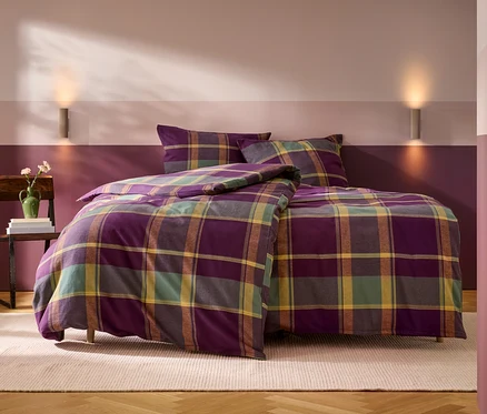 Un lit avec une parure de lit à carreaux violet, vert, jaune et marron, comprenant une couette et deux oreillers. À gauche, une chaise en bois supporte une pile de livres et un vase vert avec des fleurs. Le mur derrière le lit est peint en deux bandes horizontales, violet foncé en bas et rose-violet clair en haut, avec deux appliques murales. Un tapis clair est posé sur le parquet en bois.
