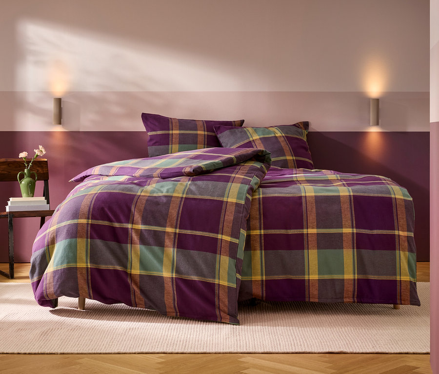 Un lit avec une parure de lit à carreaux violet, vert, jaune et marron, comprenant une couette et deux oreillers. À gauche, une chaise en bois supporte une pile de livres et un vase vert avec des fleurs. Le mur derrière le lit est peint en deux bandes horizontales, violet foncé en bas et rose-violet clair en haut, avec deux appliques murales. Un tapis clair est posé sur le parquet en bois.