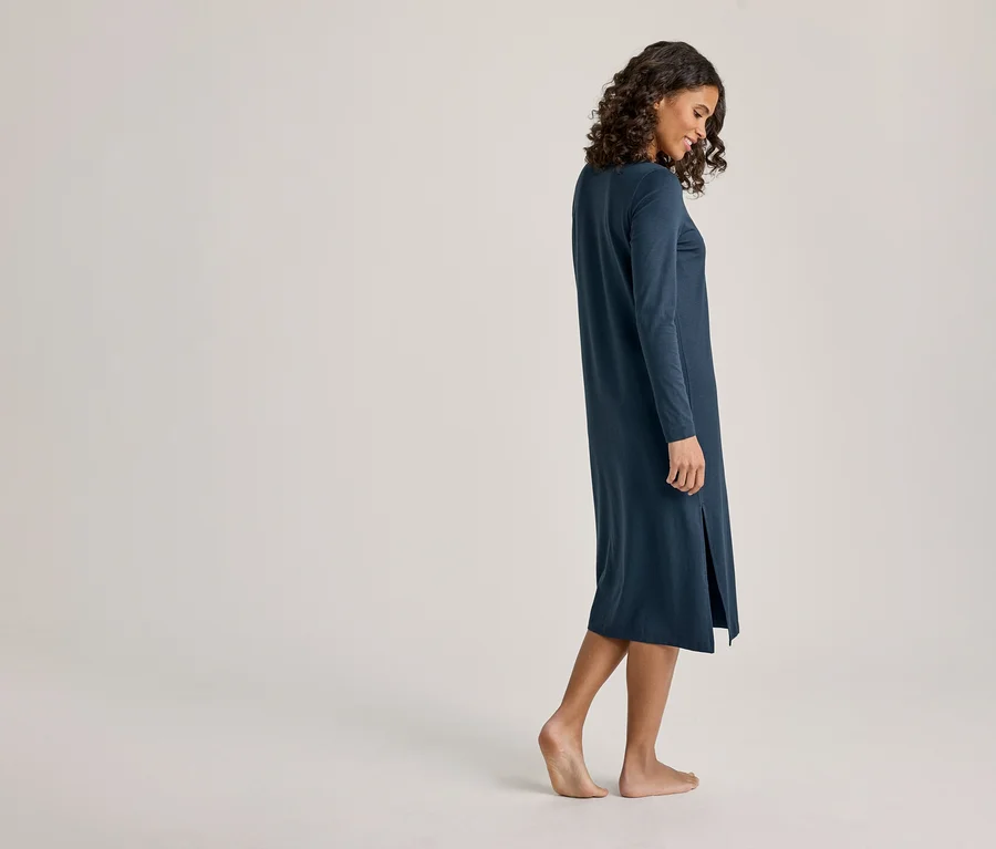 Une femme aux cheveux bouclés foncés est de profil, regardant vers le bas. Elle porte une robe midi bleu foncé à manches longues avec une fente latérale et est pieds nus.