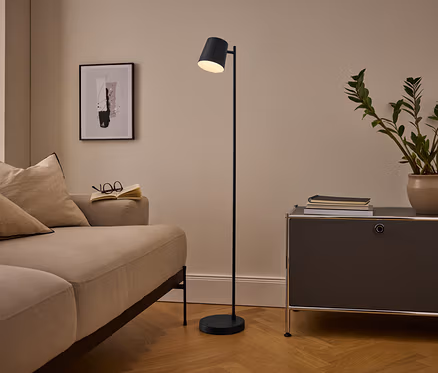 Salon confortable avec un canapé beige, un lampadaire noir et un meuble gris foncé. Un livre ouvert et des lunettes sont posés sur le canapé, une plante en pot et des livres sur le meuble. Un tableau abstrait encadré est accroché au mur.
