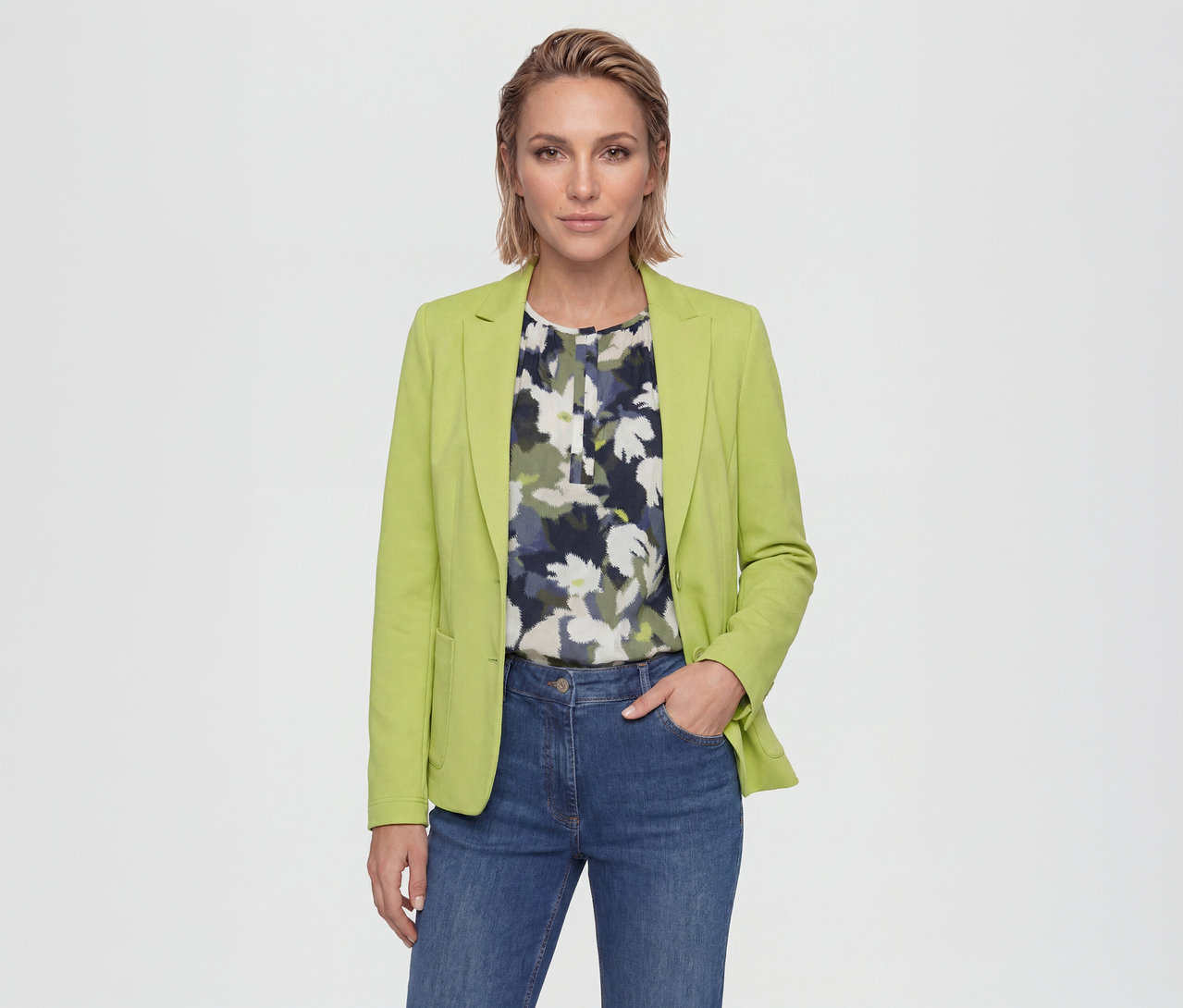 Femme portant un blazer vert citron sur un chemisier fleuri et un jean bleu, debout, une main dans la poche, regardant la caméra.
