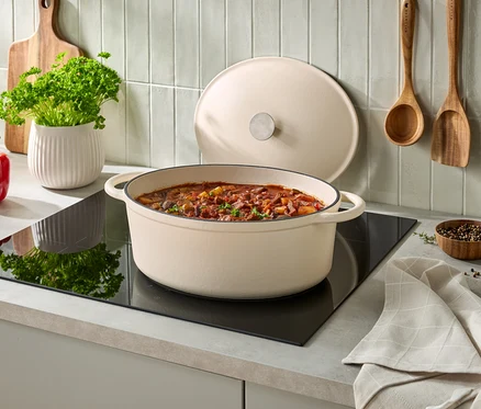 Cocotte ovale couleur crème remplie de ragoût sur une plaque à induction noire, son couvercle appuyé derrière. À gauche, un poivron rouge, un pot blanc de persil et une planche à découper en bois. Deux ustensiles en bois sont accrochés au mur carrelé, avec un petit bol de grains de poivre et un torchon plié à droite.