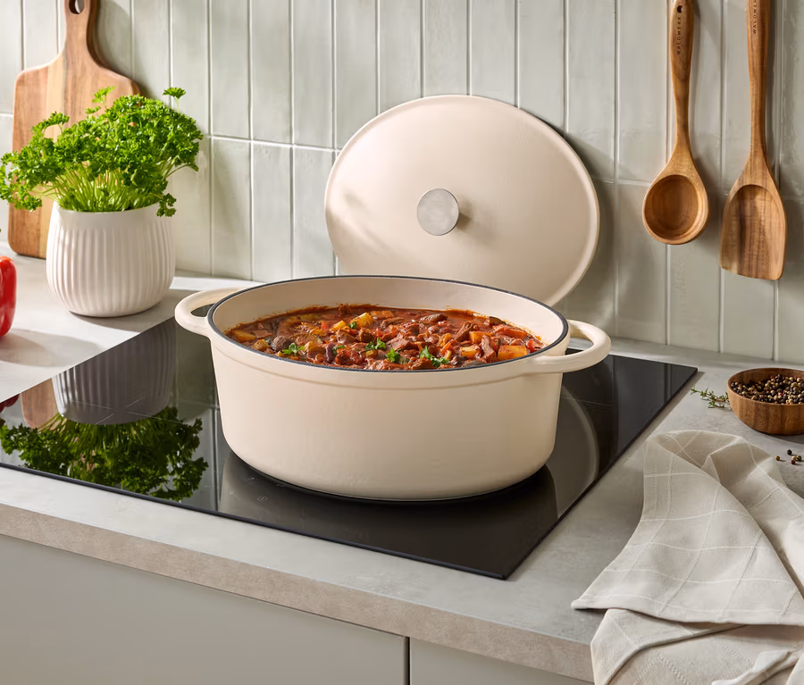 Cocotte ovale couleur crème remplie de ragoût sur une plaque à induction noire, son couvercle appuyé derrière. À gauche, un poivron rouge, un pot blanc de persil et une planche à découper en bois. Deux ustensiles en bois sont accrochés au mur carrelé, avec un petit bol de grains de poivre et un torchon plié à droite.