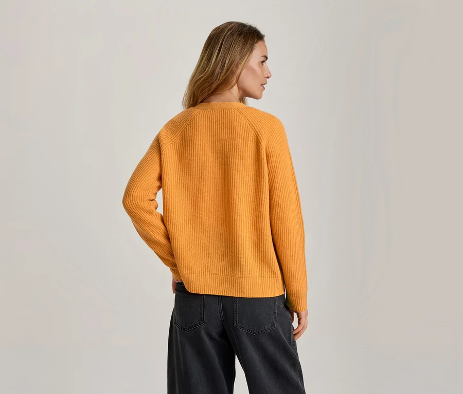 Femme vue de dos, légèrement tournée vers la droite, portant un pull côtelé orange et un jean large gris foncé.