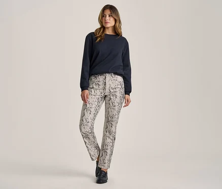 Femme debout, portant un haut à manches longues bleu foncé, un pantalon gris clair à motif peau de serpent noir et des chaussures noires.